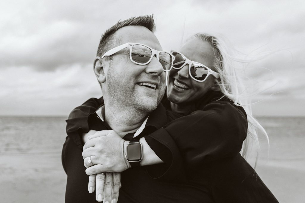 Lachendes Paar mit Sonnenbrillen bei einem Verlobungsshooting am Elbstrand in Hamburg.